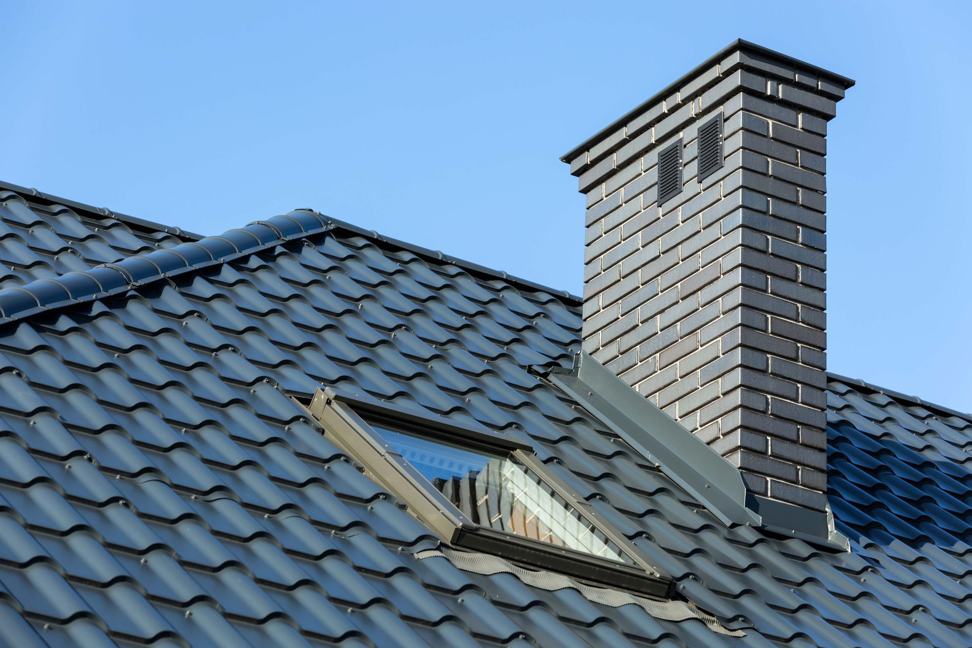 Black tiled roof with chimney and skylight against a blue sky.