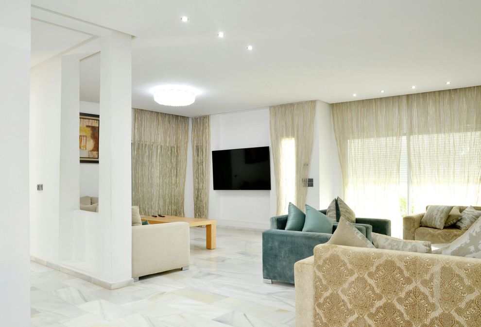 Spacious living room with large TV, neutral tones, multiple couches, and natural light from sheer curtains.