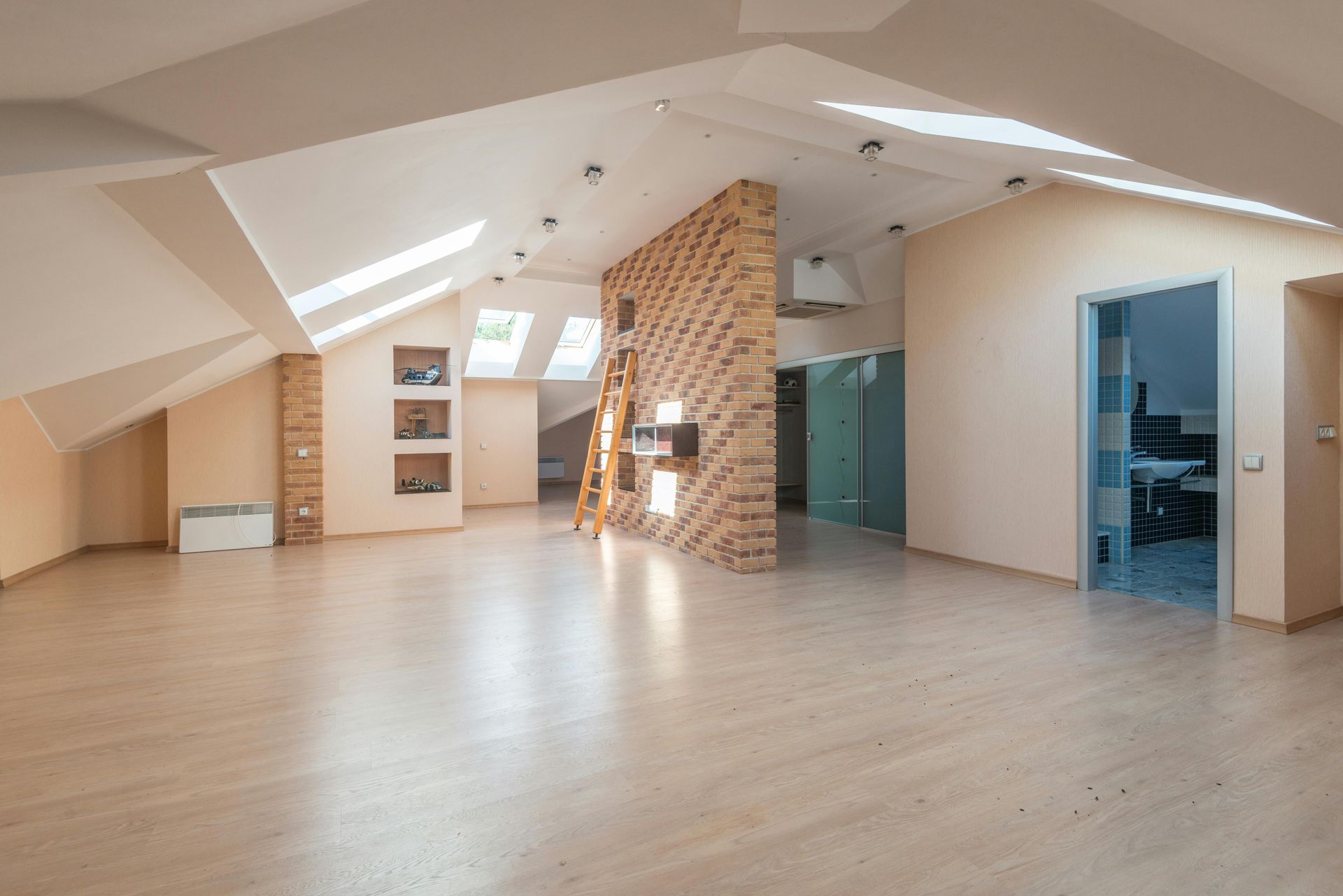 Spacious, sunlit attic room with slanted ceilings, skylights, brick accent wall, light wood floor, and doorway.