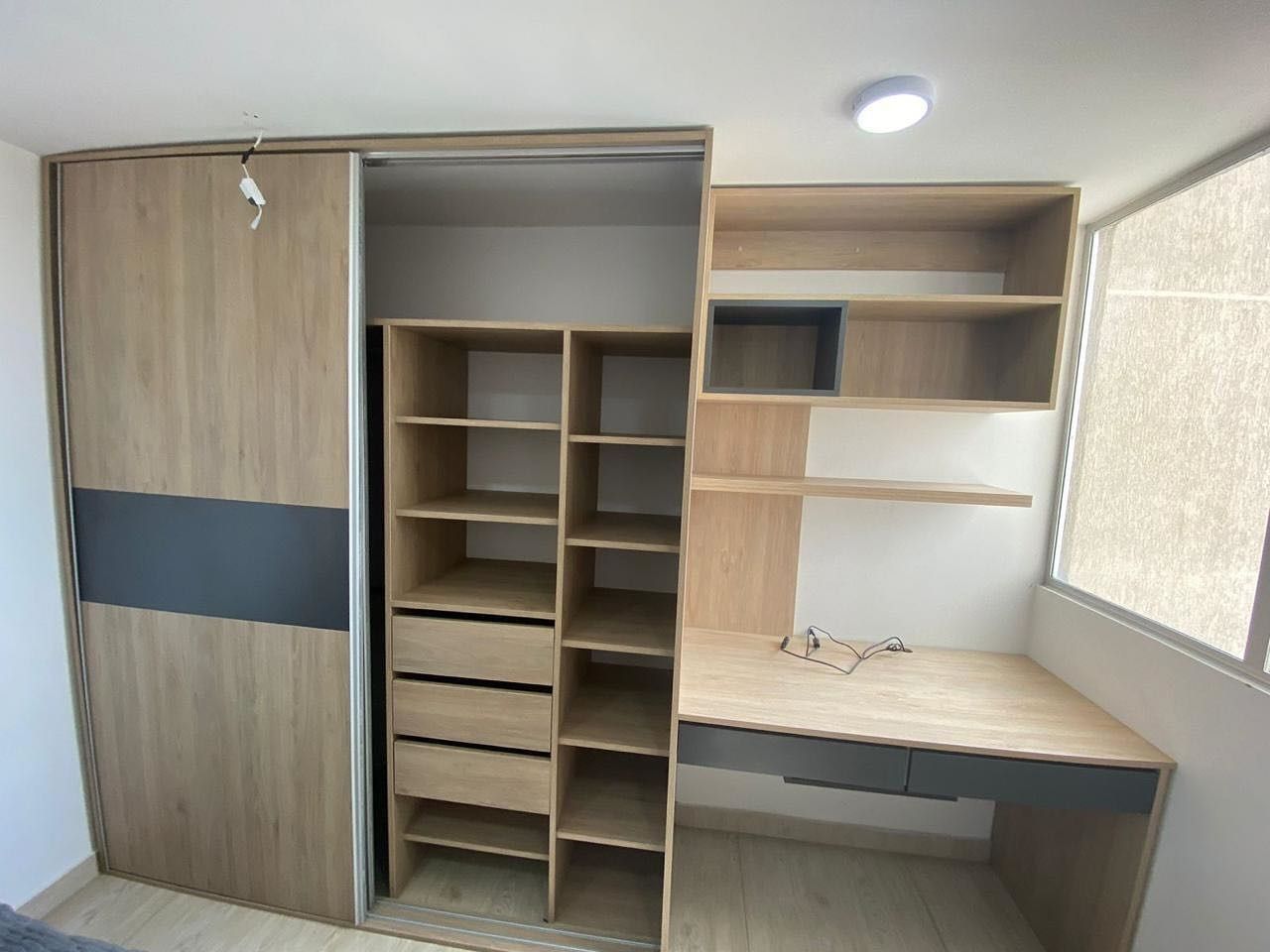 Escritorio empotrado de madera y estantería con armario, en color marrón claro y gris, junto a una ventana.