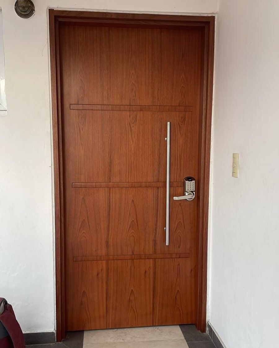 Puerta de madera con tirador vertical plateado y cerradura digital. Empotrada en una pared blanca.