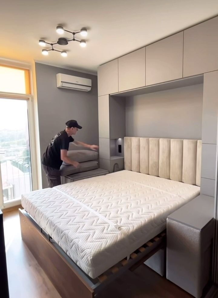 Hombre colocando almohadas sobre una cama en un dormitorio moderno con cabecera y armarios incorporados; colchón de color claro.