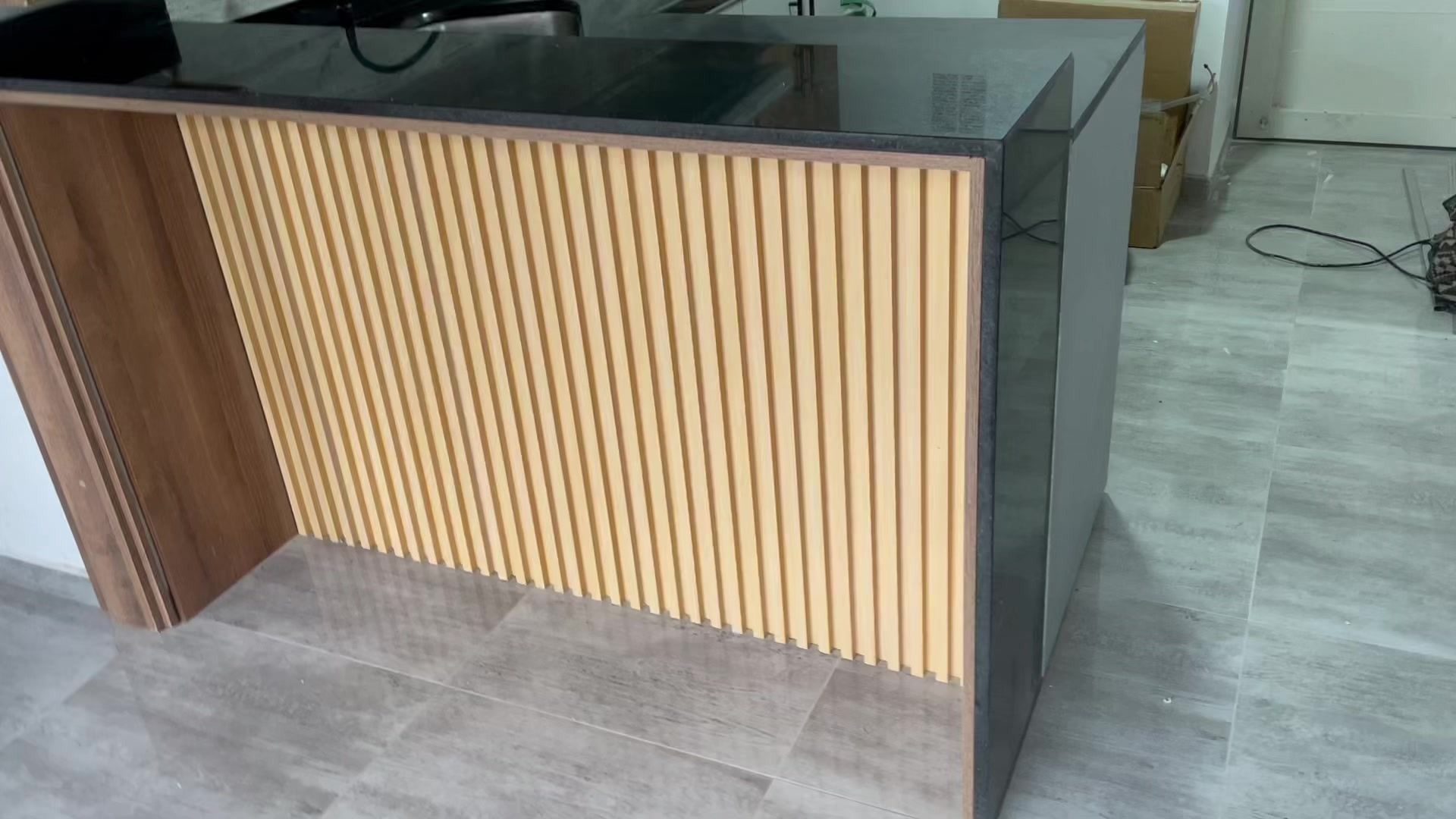 Mostrador de recepción con paneles de madera y cristal, lamas verticales en color beige claro, encimera y lateral en color gris.