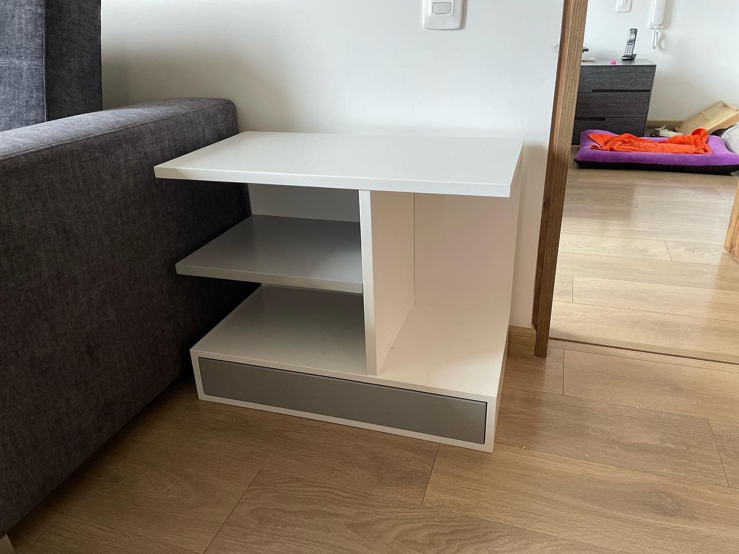 Mesa auxiliar blanca con dos estantes abiertos y un cajón gris junto a un sofá gris.