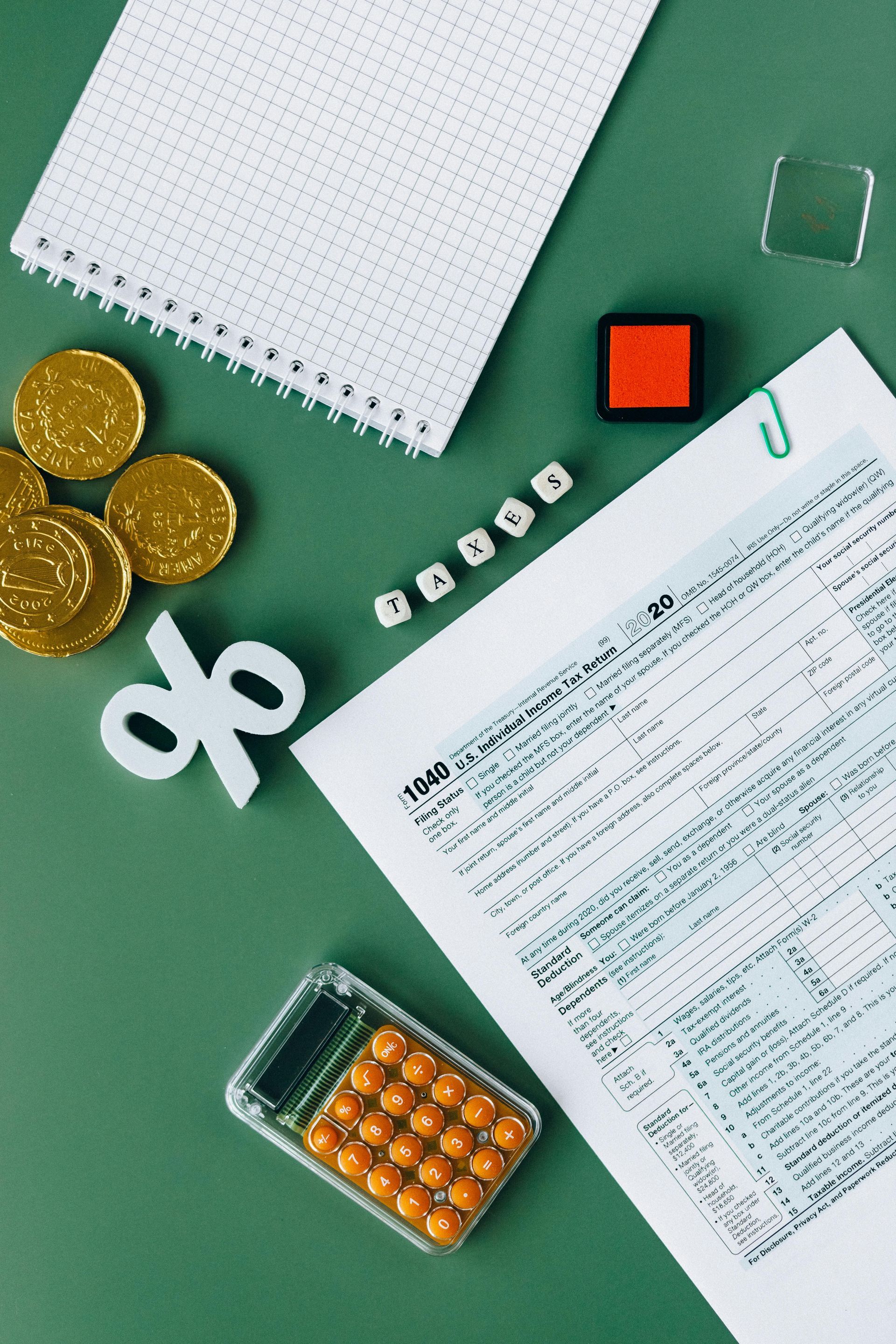 A flat-lay view of financial tools: coins, a percentage sign, a calculator, a notepad, and a tax form on a green surface.