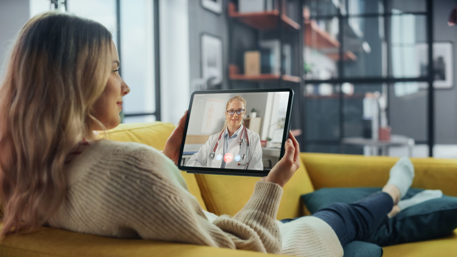 A woman is sitting on a couch holding a tablet with a doctor on it.