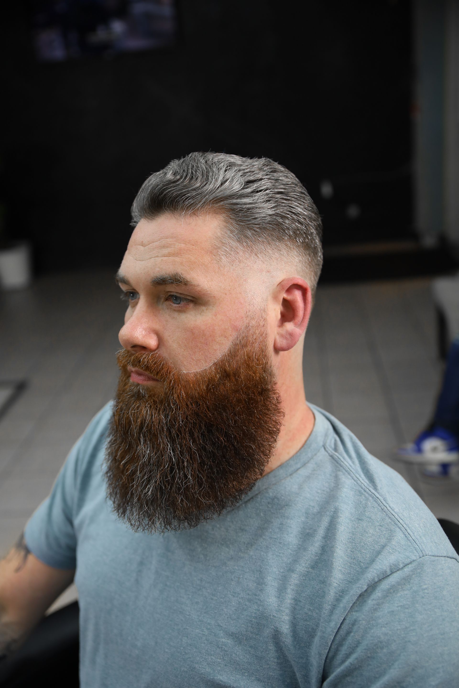 Man with a thick, reddish-brown beard and faded gray hair. He wears a blue shirt and sits indoors.