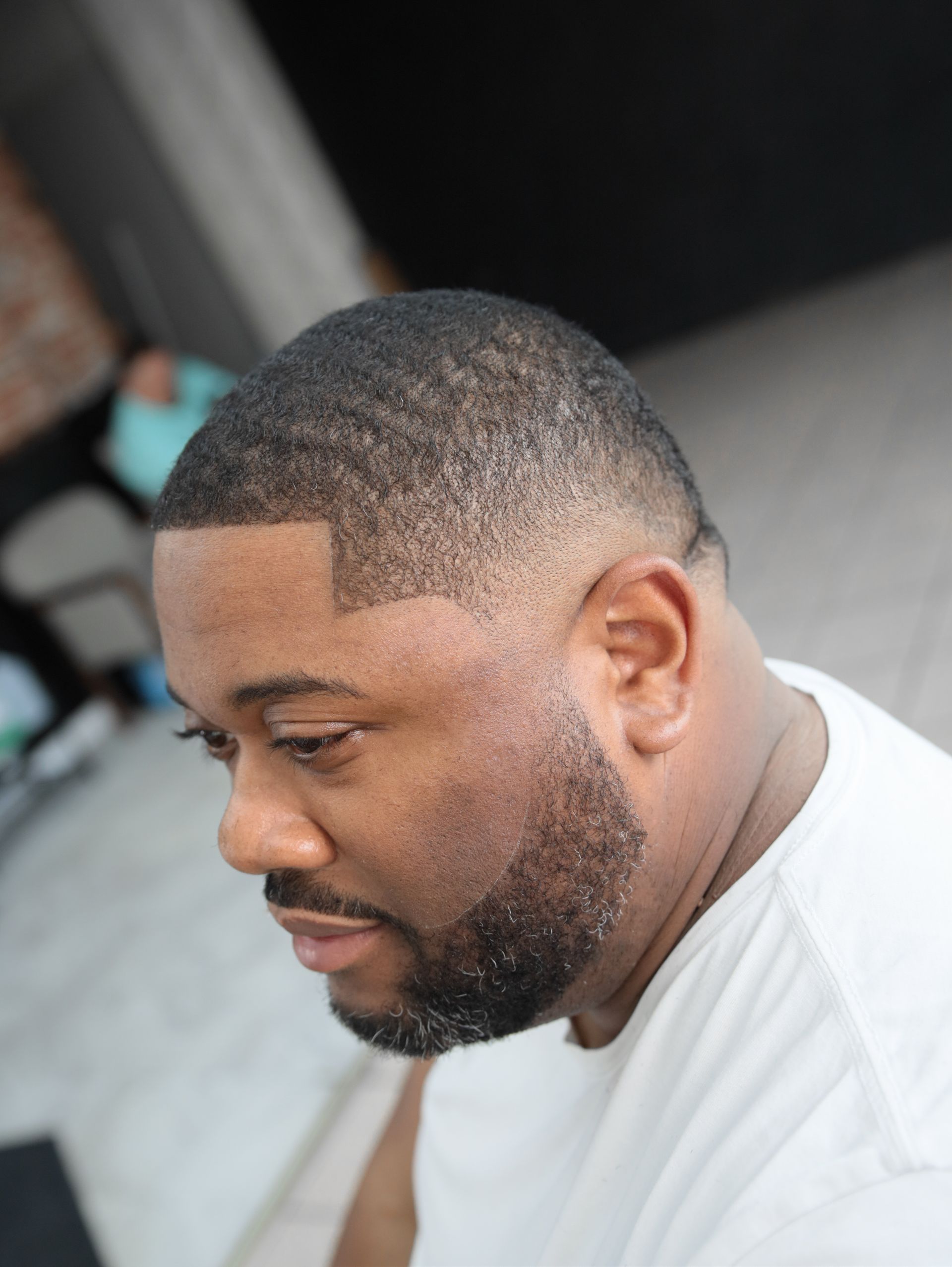 Man with dark skin, wearing a white shirt, looking down. Faded haircut, trimmed beard.