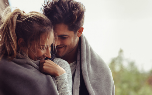Couple wrapped in a gray blanket, smiling, standing outdoors.