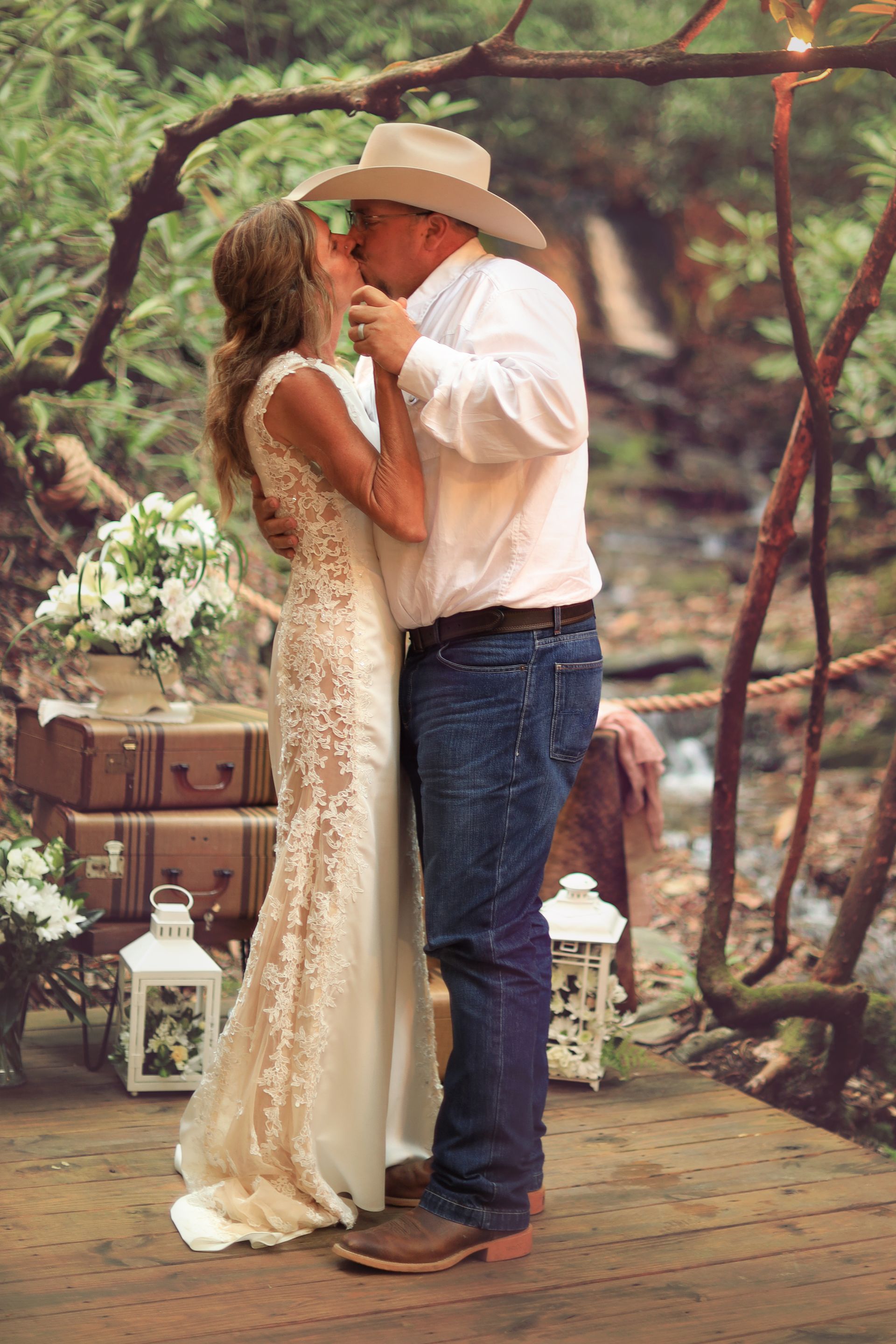 Bride and groom kissing outdoors at wedding ceremony, on a wooden bridge, surrounded by foliage and decorations.