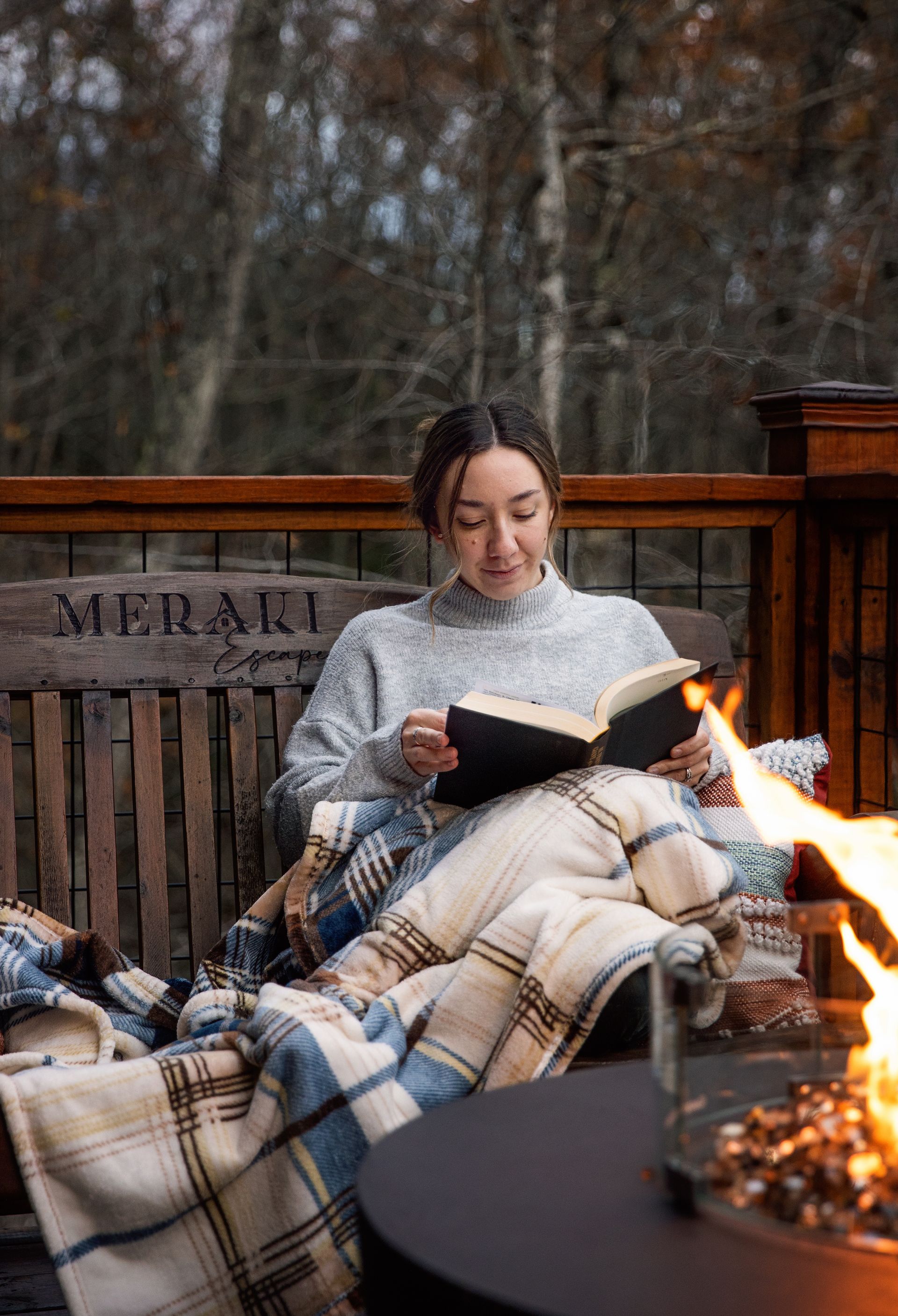 A woman is sitting on a bench reading a book next to a fire pit.