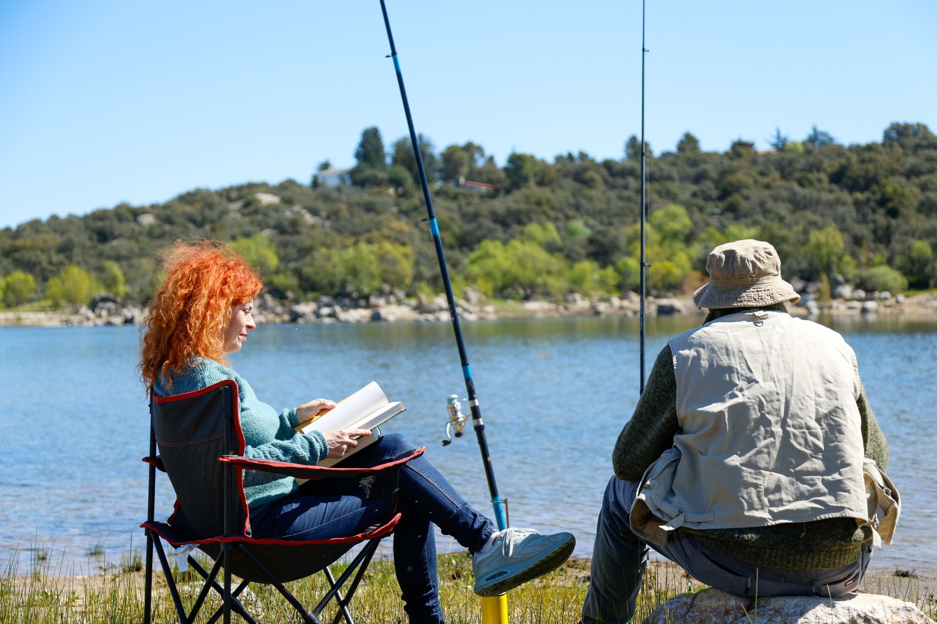 A man and a woman are sitting on the shore of a lake with fishing rods.