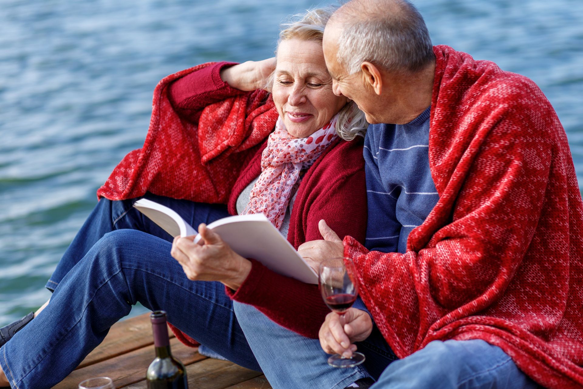 A man and a woman are sitting on a dock reading a book and drinking wine.