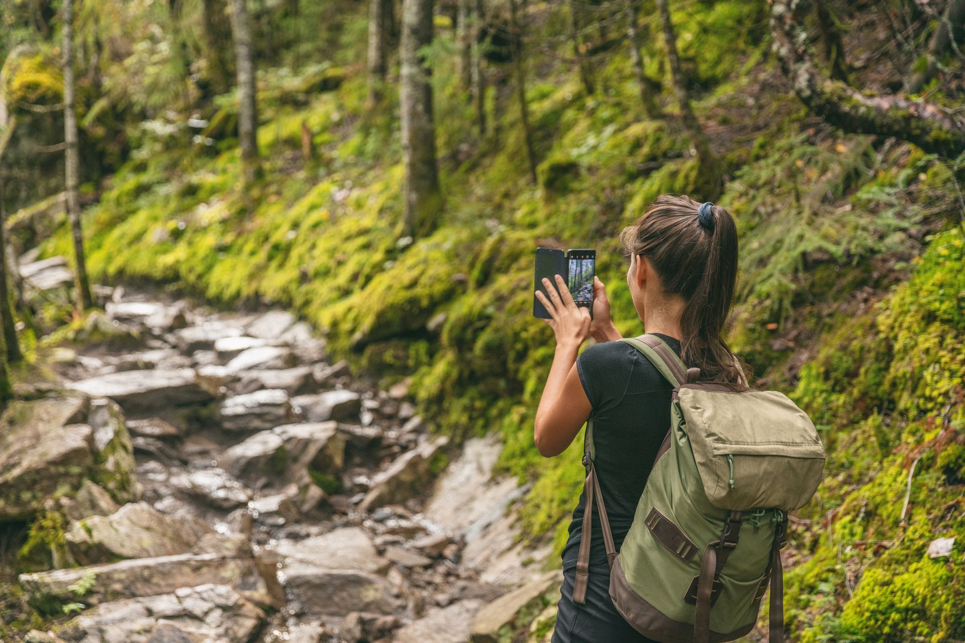 A woman with a backpack is taking a picture of a stream in the woods.