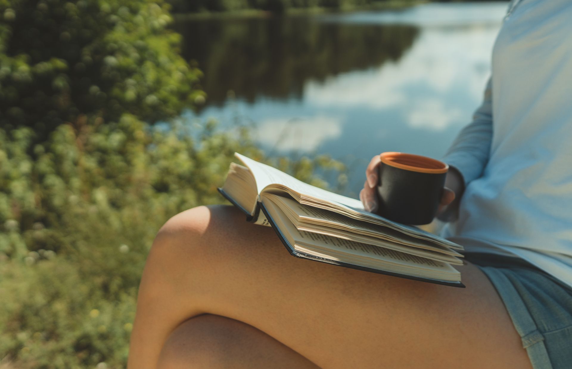 A woman is sitting on the grass reading a book and holding a cup of coffee.