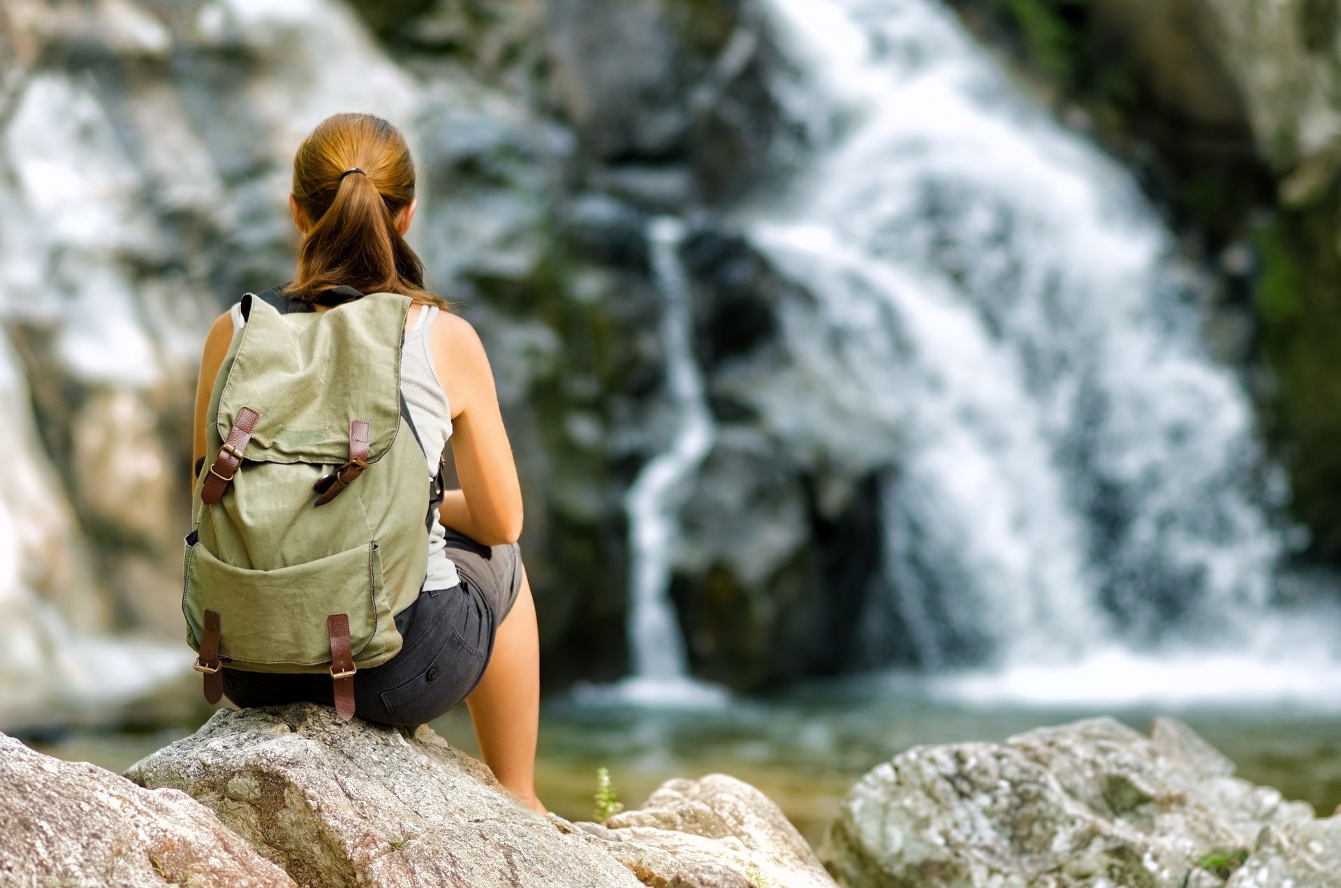 A woman with a backpack is sitting on a rock near a waterfall.