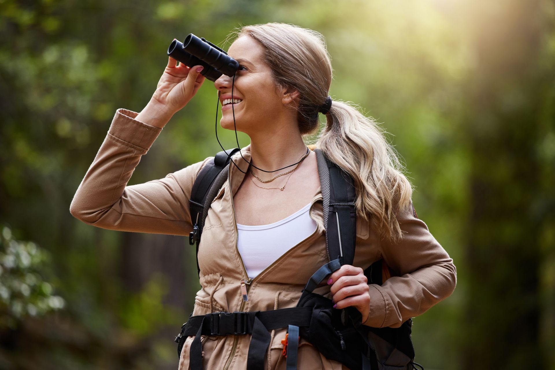 A woman with a backpack is looking through binoculars in the woods.