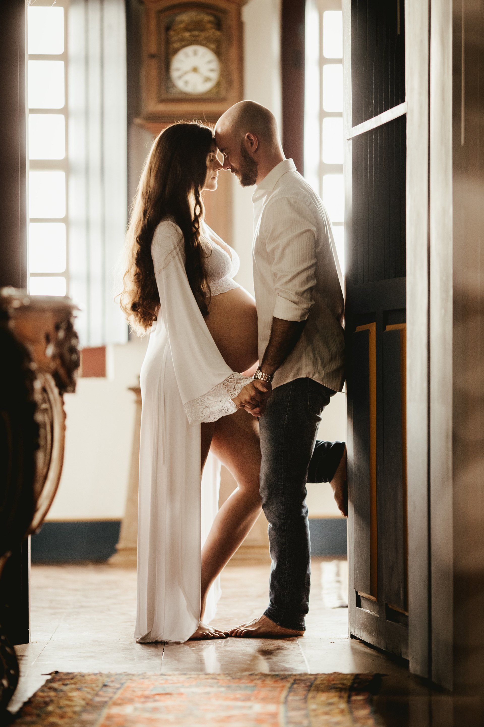 Pregnant woman and partner embrace, holding hands, in a well-lit room with a clock and a dark cabinet.