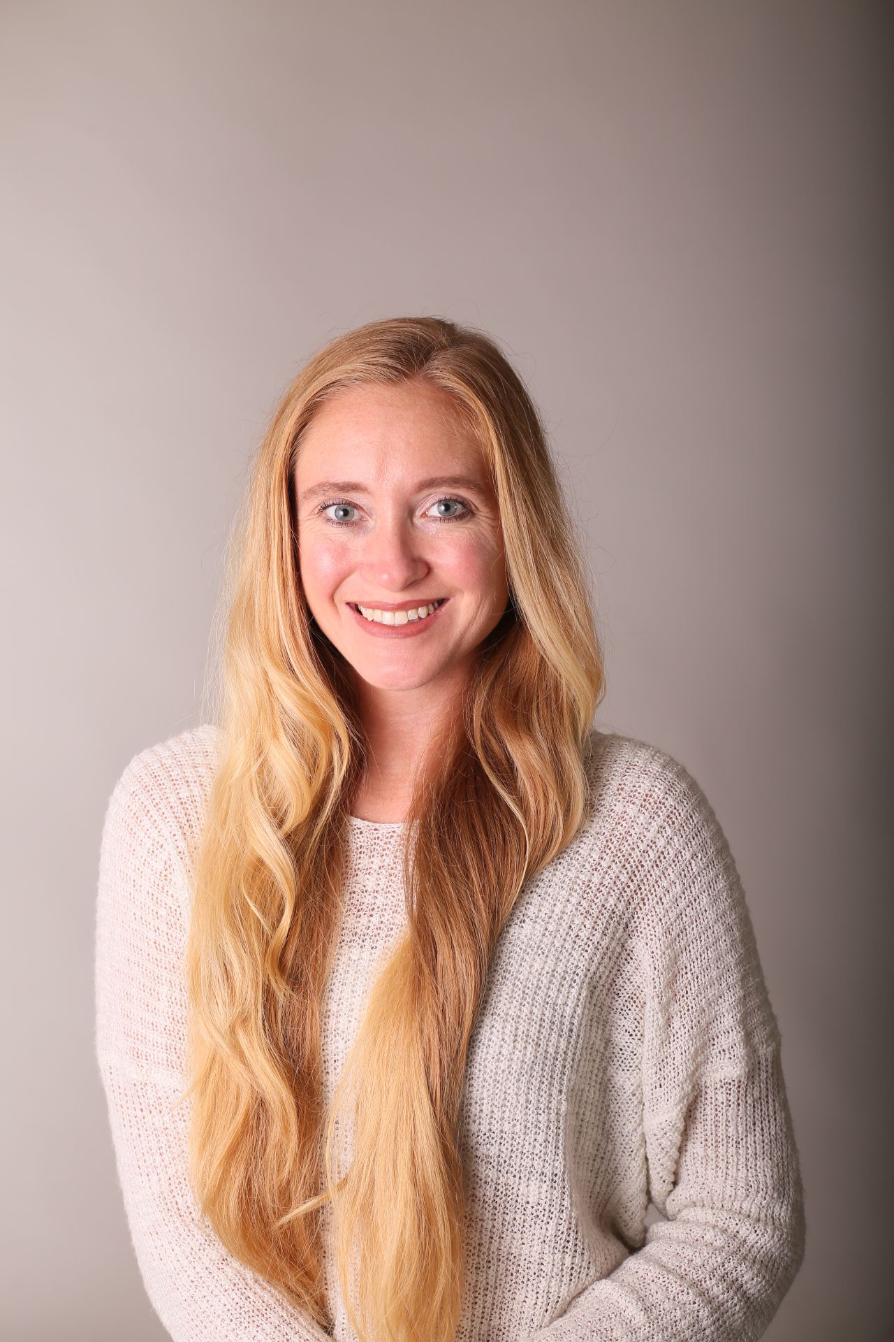 A woman with long blonde hair is wearing a white sweater and smiling.