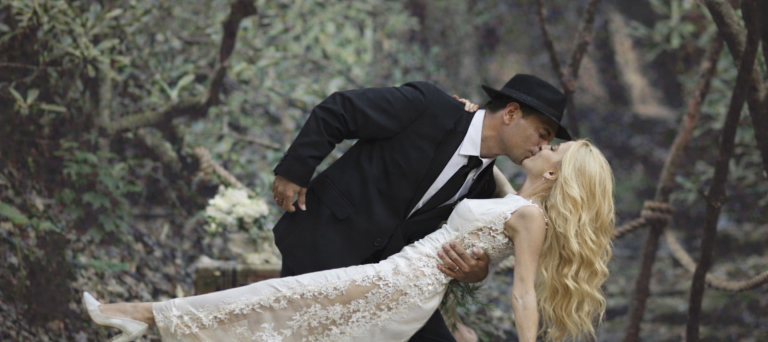 Bride and groom on wooden bridge, waterfall backdrop, romantic outdoor setting.