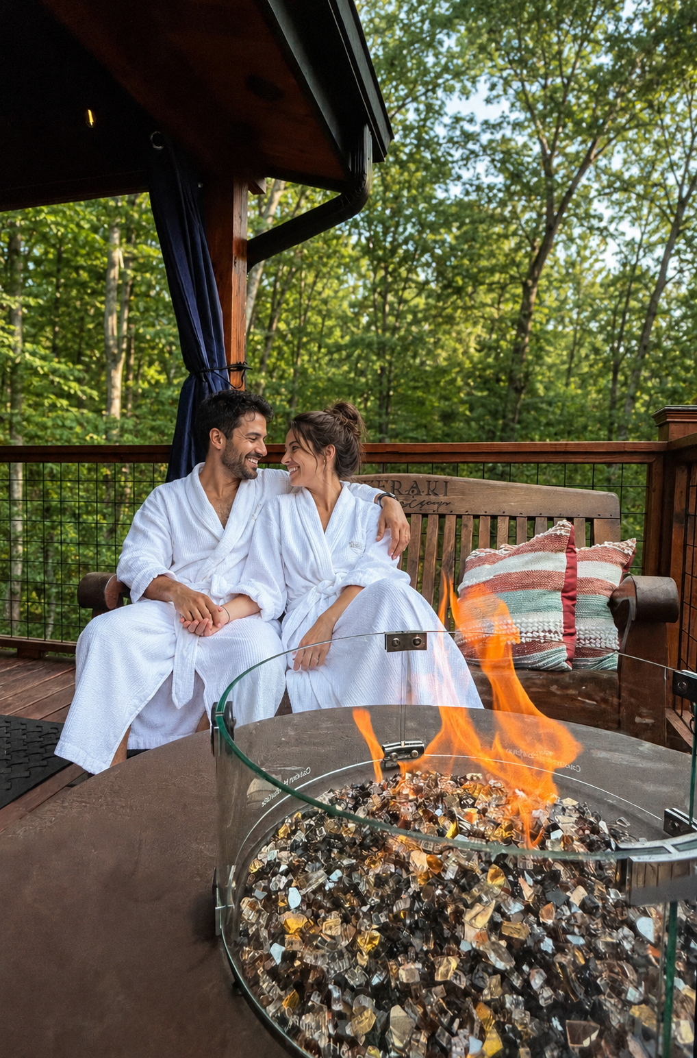 Couple in white robes sitting by a fire pit on a wooden deck surrounded by trees