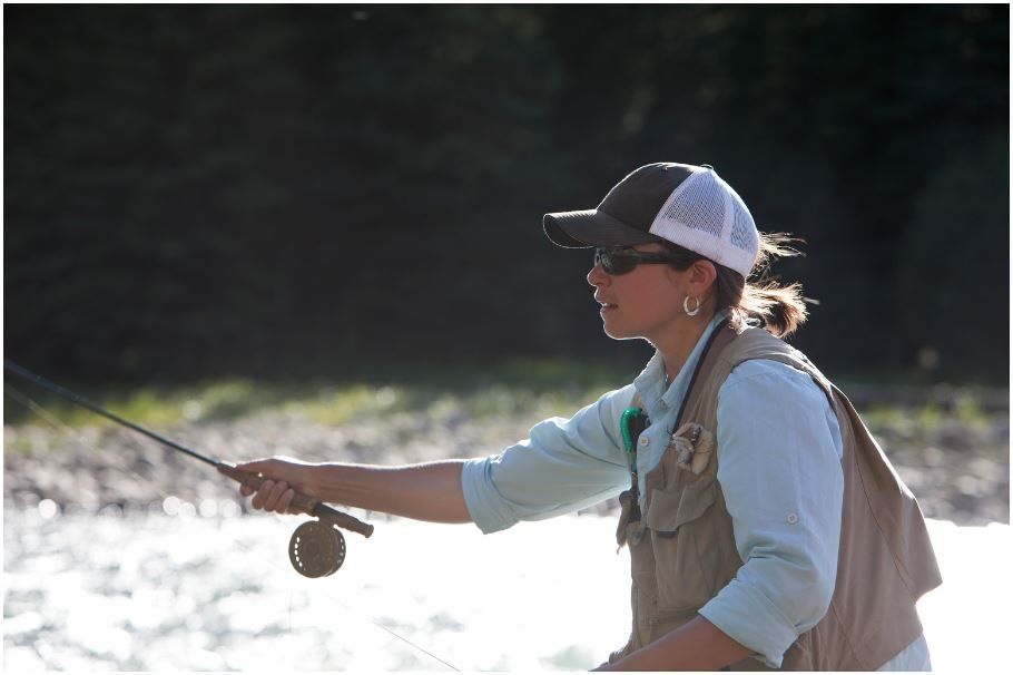 A woman is fishing in a river wearing a hat and vest