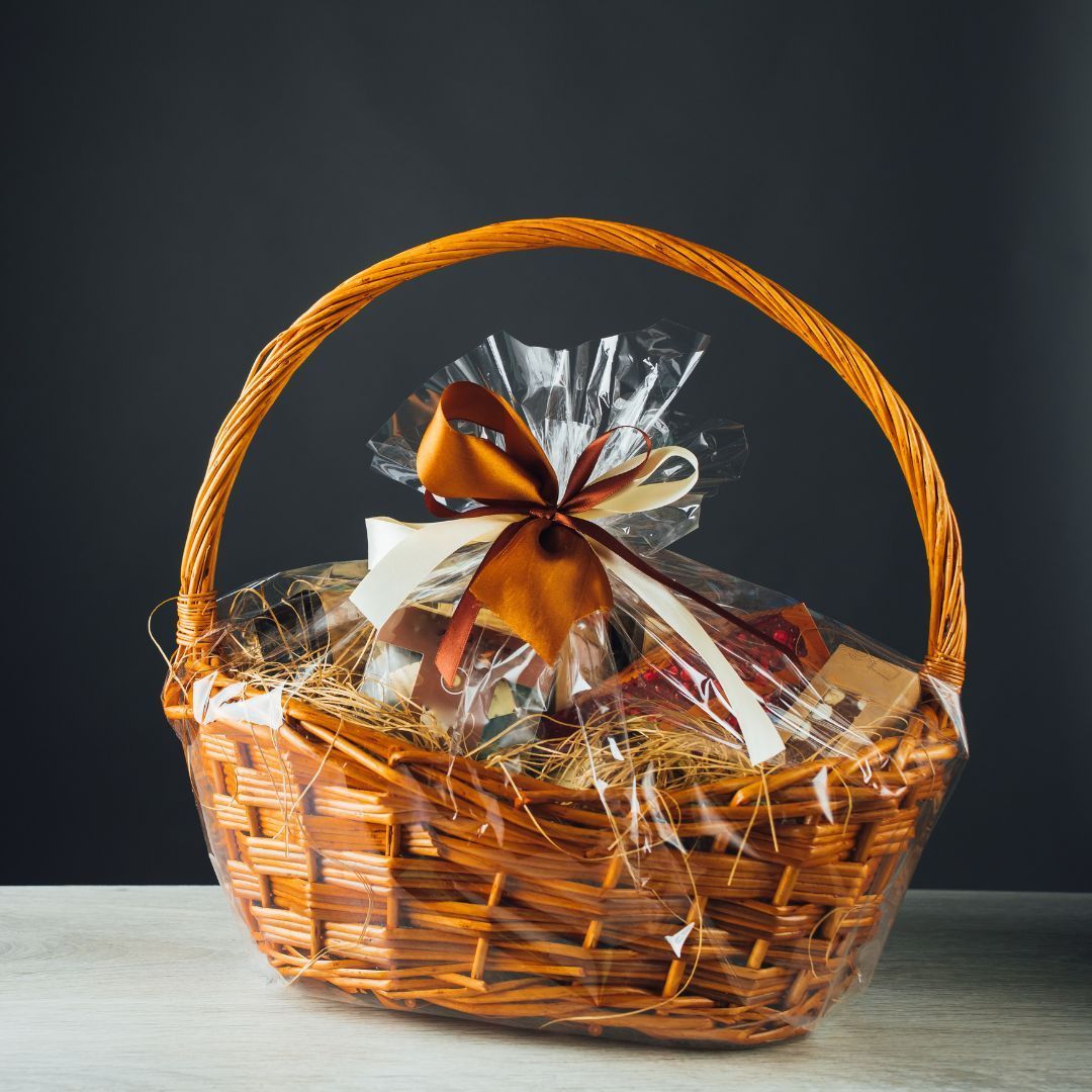Wicker gift basket wrapped in clear cellophane, tied with a brown ribbon, on a light surface against a dark background.