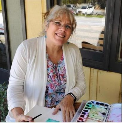 A woman is smiling while sitting at a table with watercolors
