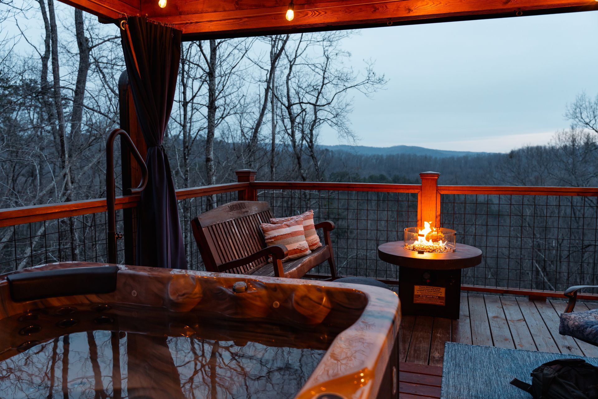 Cozy covered deck at dusk with wooden chairs, lit fire pit, and snowy forest view beyond the railing