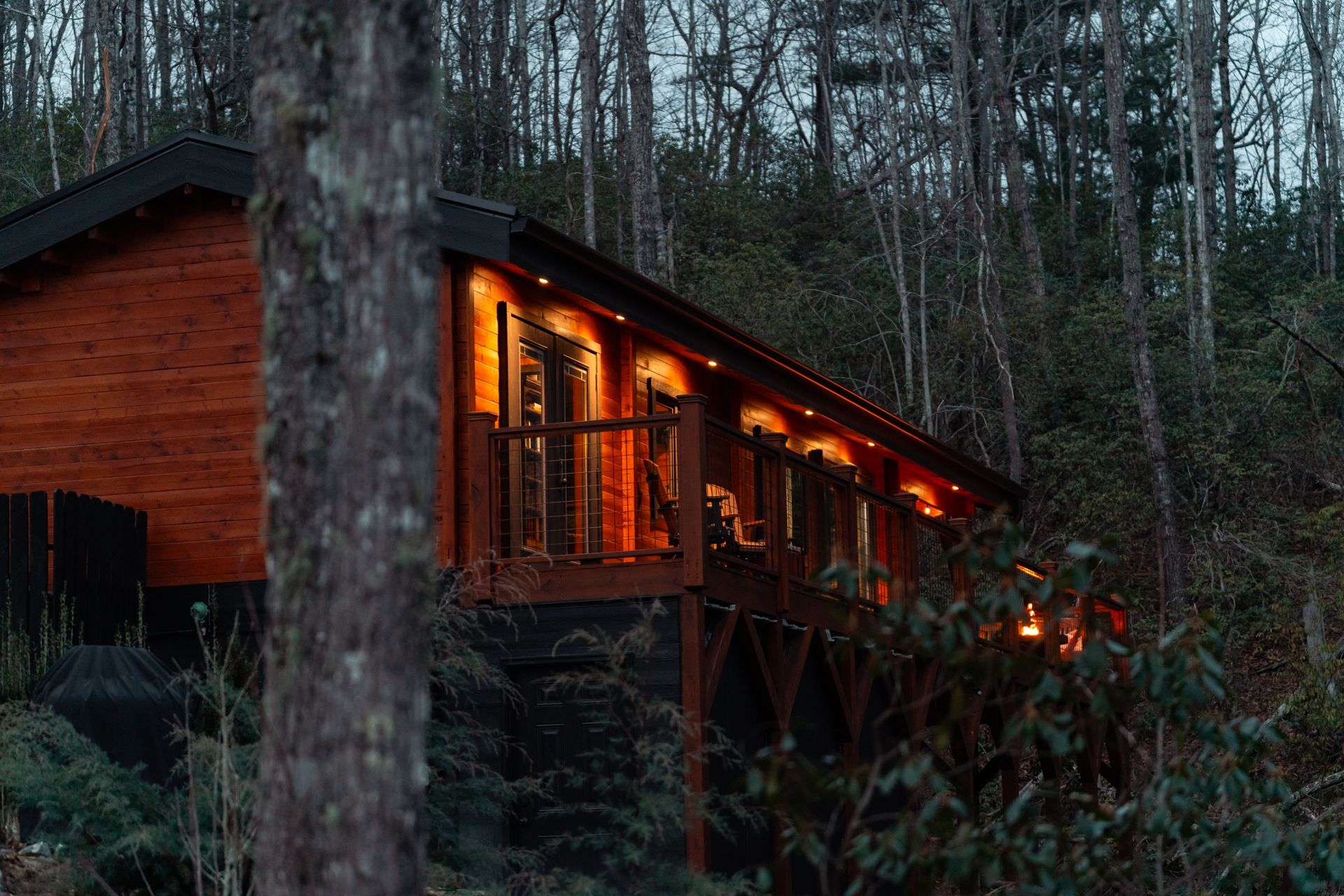 A warm, golden-lit wooden cabin with a deck nestled among trees in a dim, forest setting at twilight.