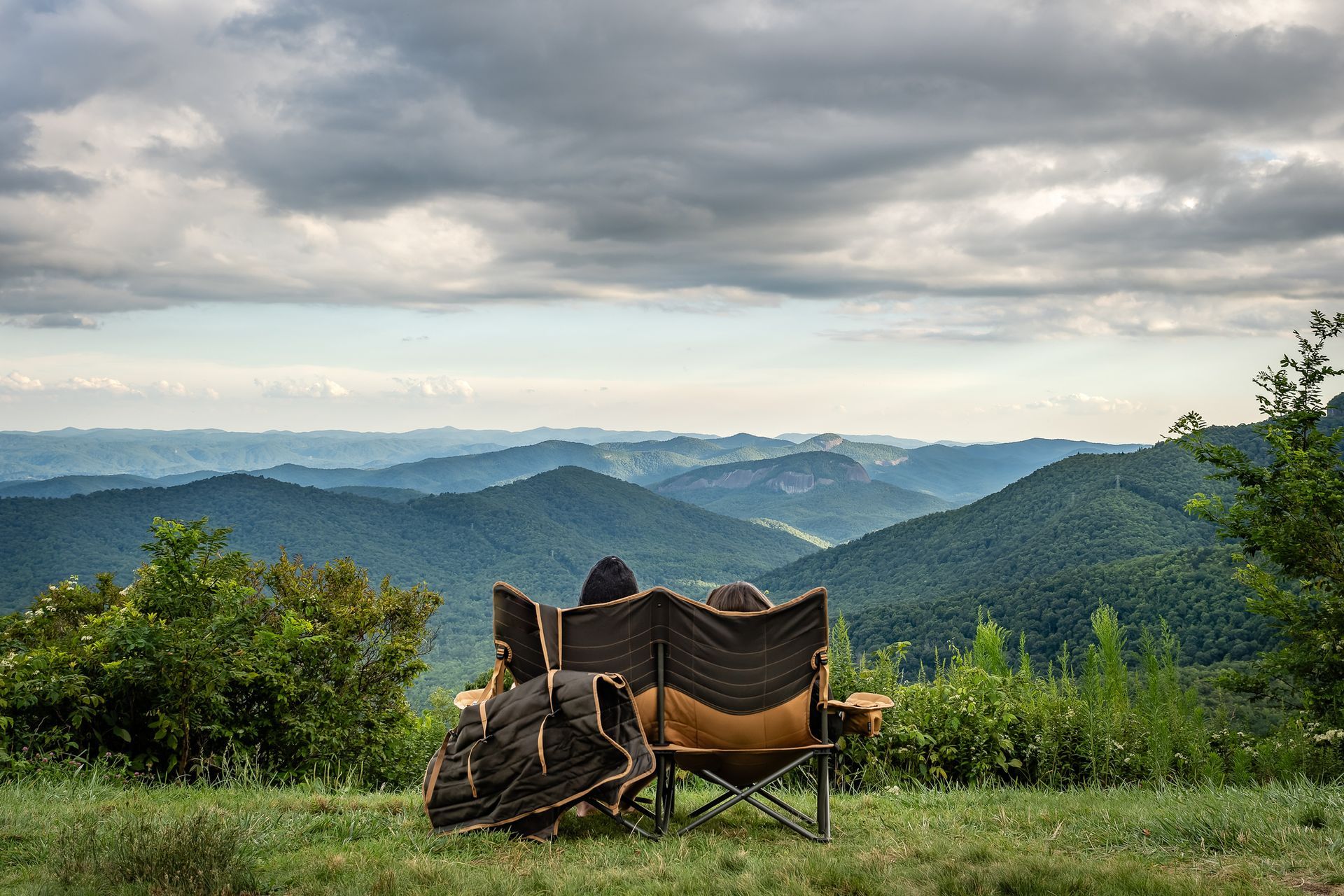 Two people in camp chairs overlooking a mountain range under a cloudy sky.