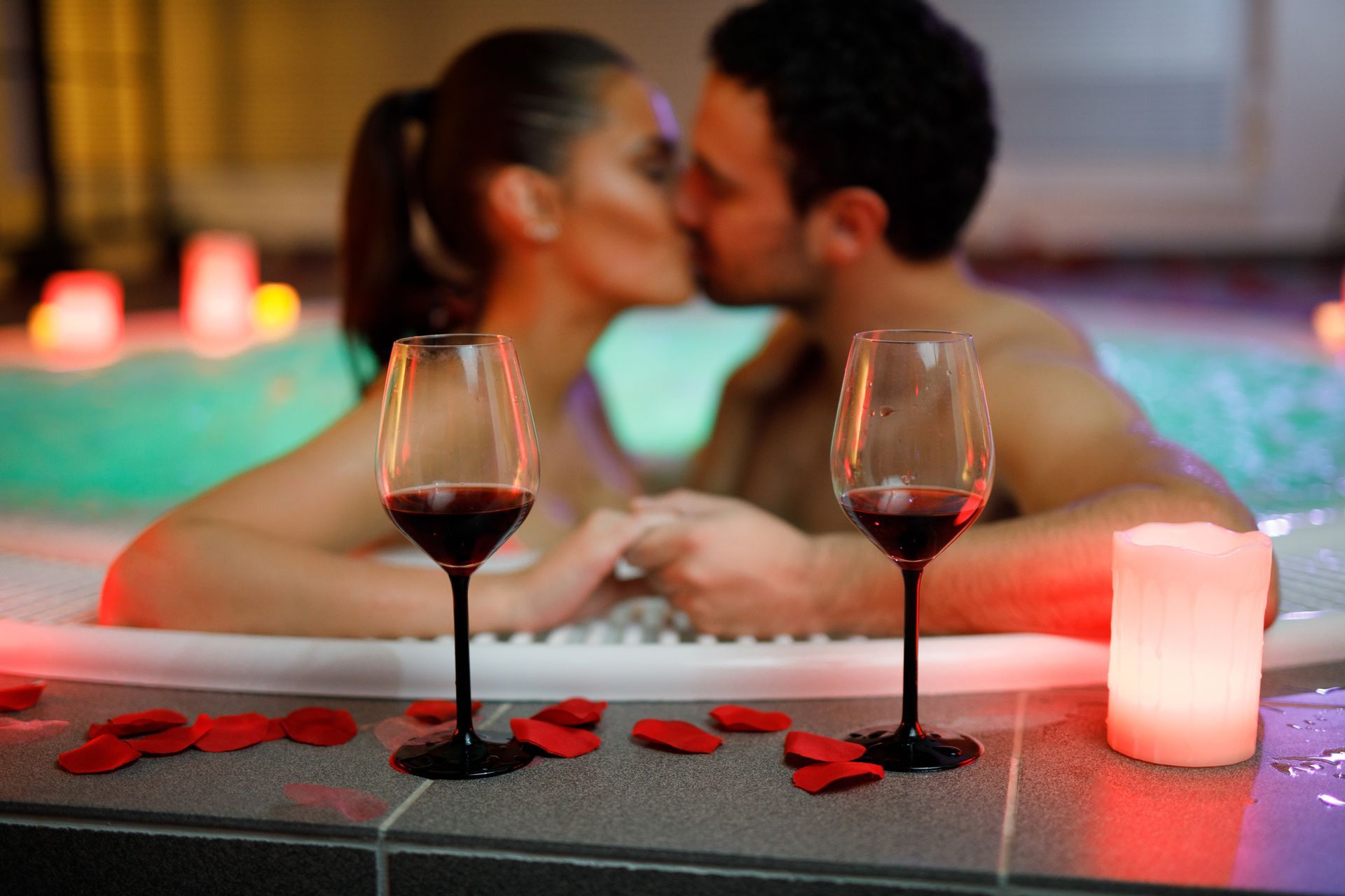 A man and a woman are kissing in a hot tub with wine glasses and candles.