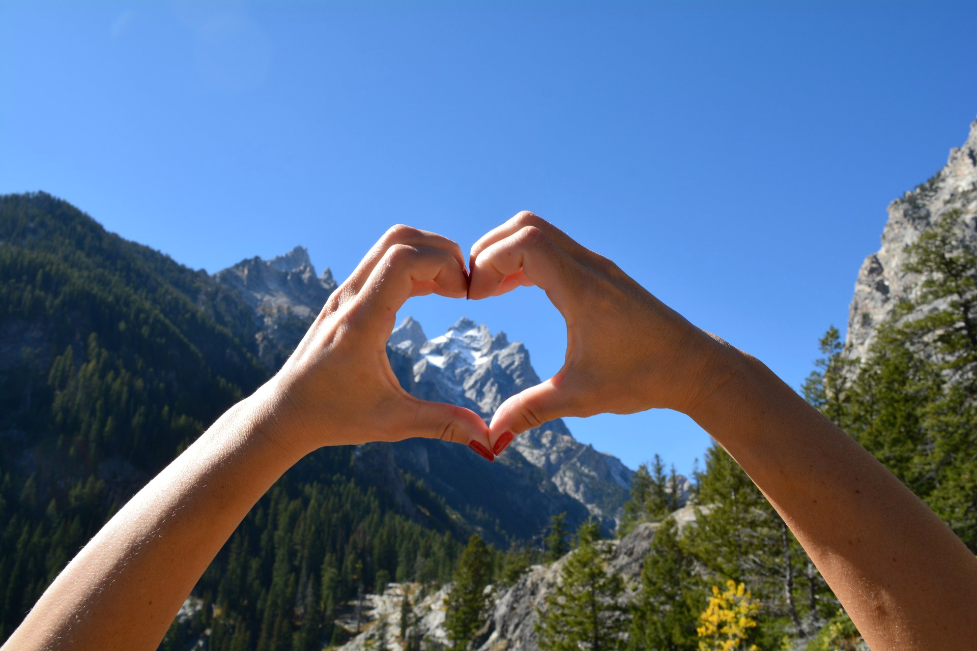 A person is making a heart shape with their hands in front of a mountain.