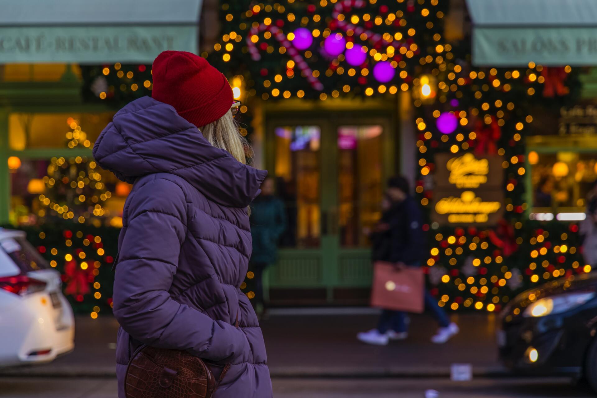 A woman in a purple jacket and red hat is standing in front of a christmas tree.