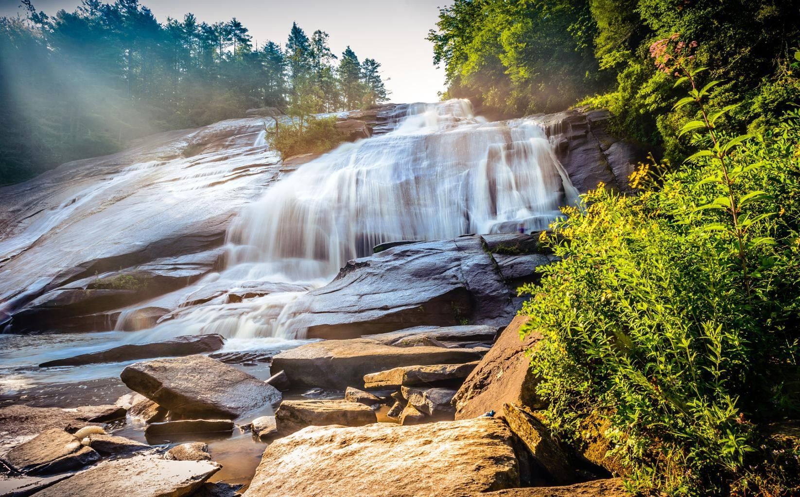 Water cascades down a sunlit, rocky slope surrounded by lush green trees, with light rays filtering through the canopy.