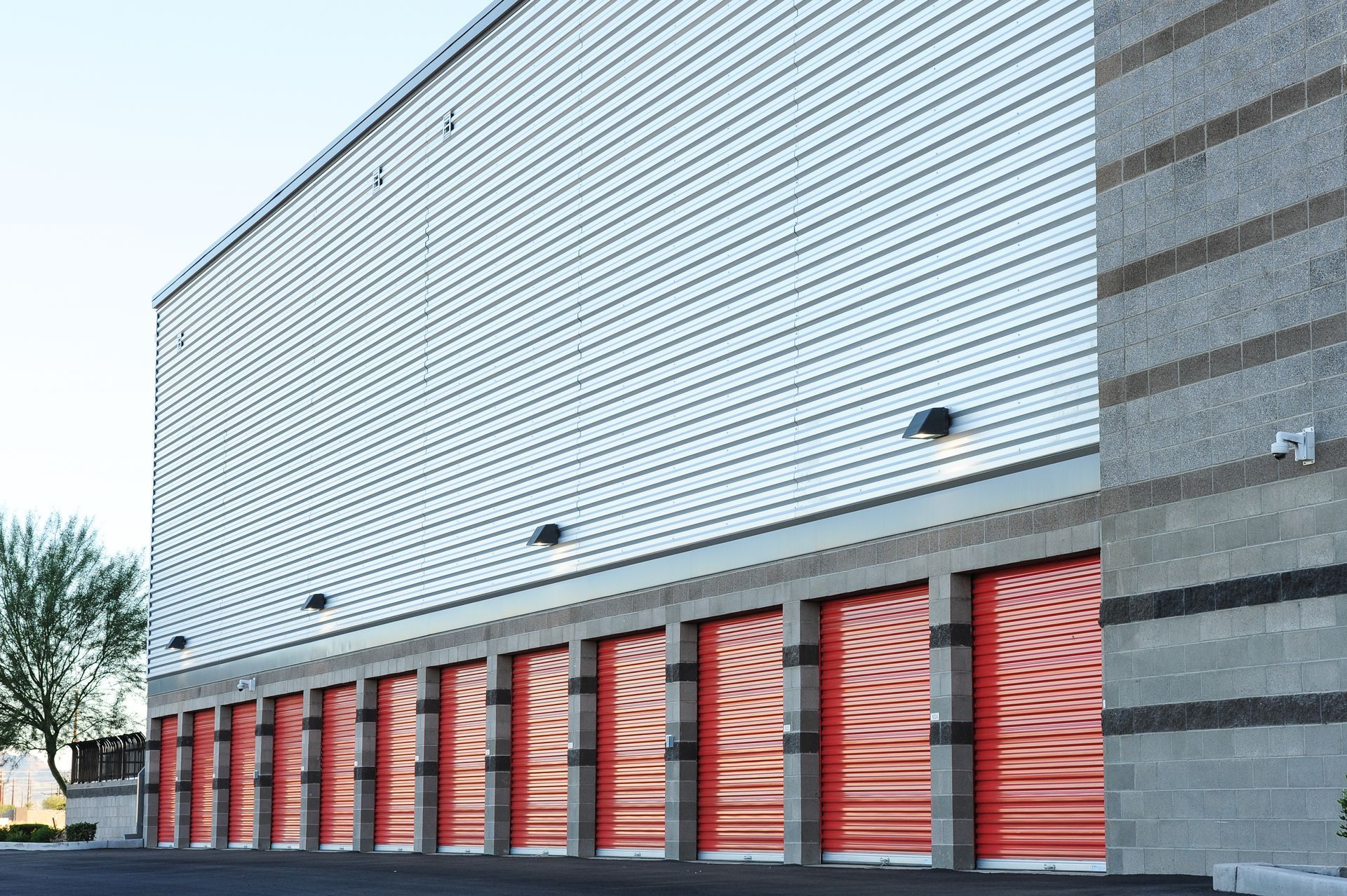 Storage units with red doors under a large white building. Concrete and corrugated metal exterior.