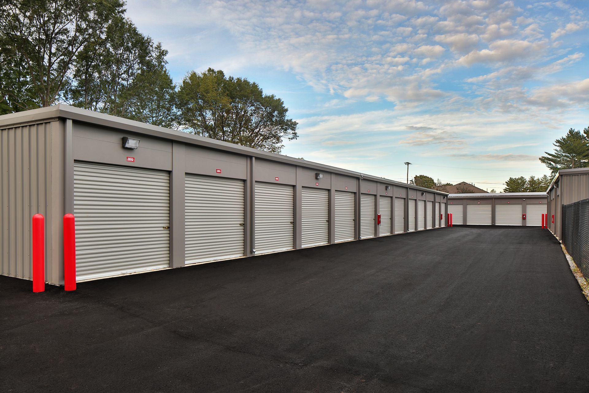 Exterior view of a self-storage facility with several grey units and a black asphalt driveway, under a cloudy blue sky.