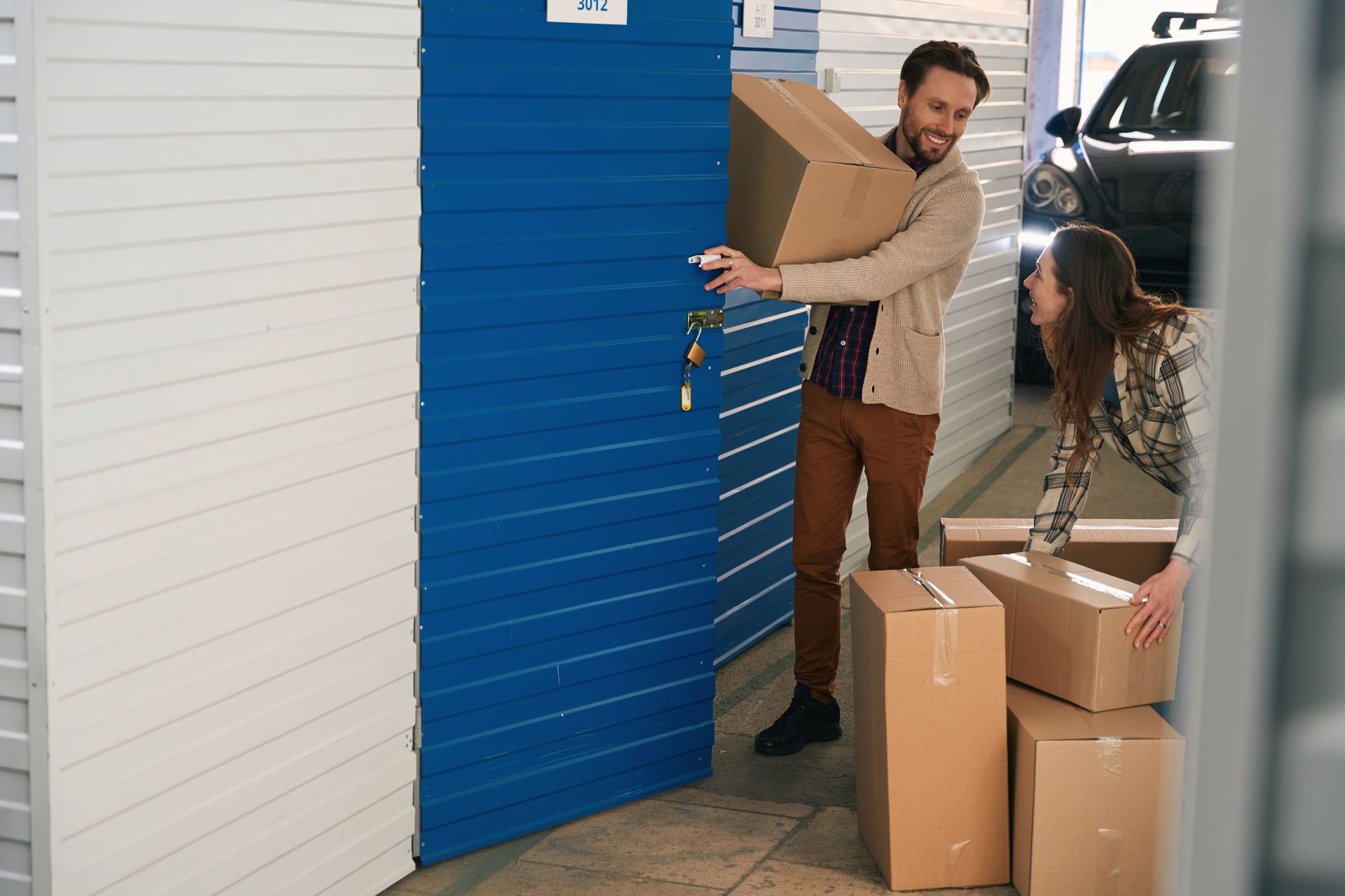 Couple loading cardboard boxes into a blue storage unit. Man holds a box, smiling.
