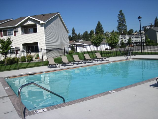 Swimming pool with lounge chairs next to a two-story apartment building under a blue sky.