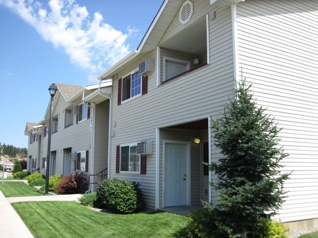 Two-story beige apartment building with maroon shutters, green lawn, and blue sky.