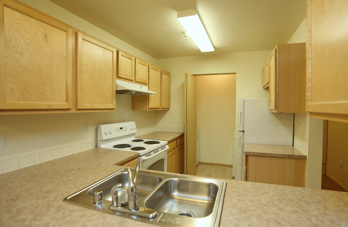 Kitchen with light wood cabinets, a stove, and a double sink. Beige countertops and a white refrigerator are visible.