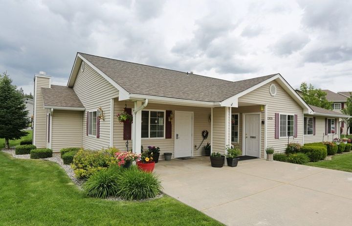 Tan, single-story homes with white doors, a shared carport, and a green lawn under a cloudy sky.