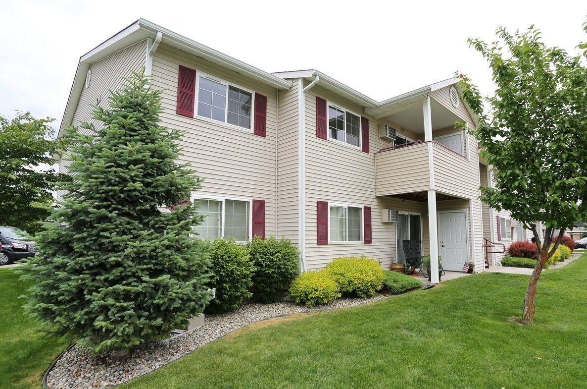 Beige two-story apartment building with red shutters, green lawn, and evergreen trees.