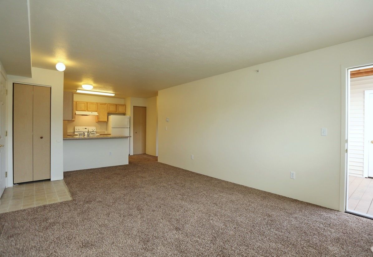 Empty apartment living room with brown carpet and off-white walls, kitchen in the background.