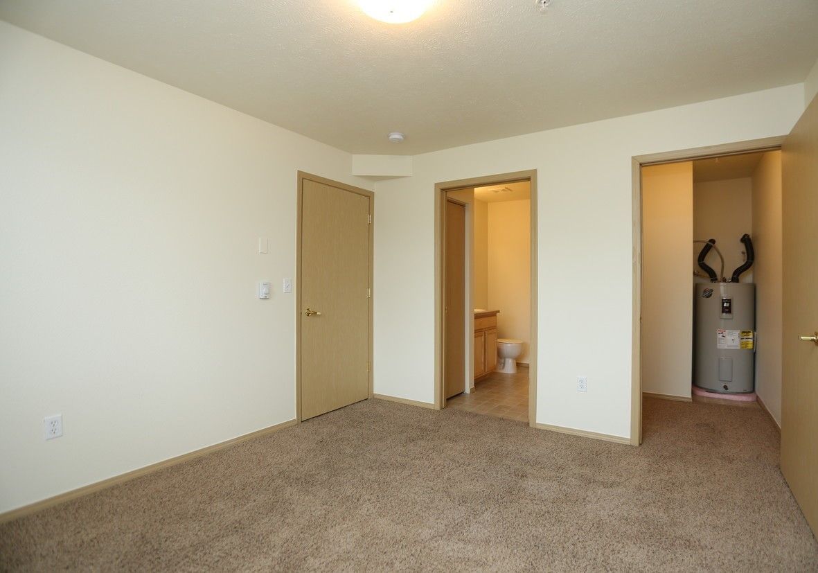 Empty room with beige walls, carpet, and three doorways, including a bathroom and water heater closet.