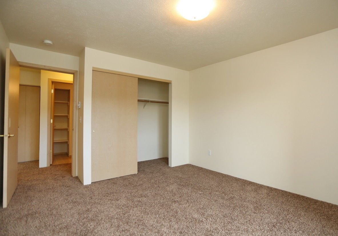 Empty room with beige walls, brown carpet, closet, and open doorway to a hallway.