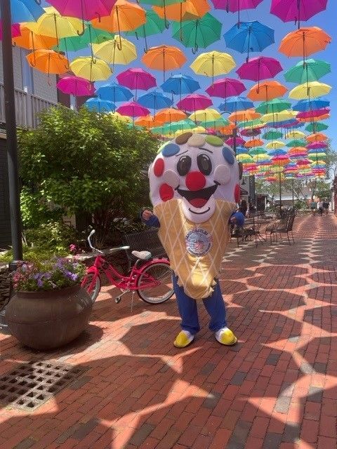 An ice cream cone mascot is standing in front of a row of colorful umbrellas