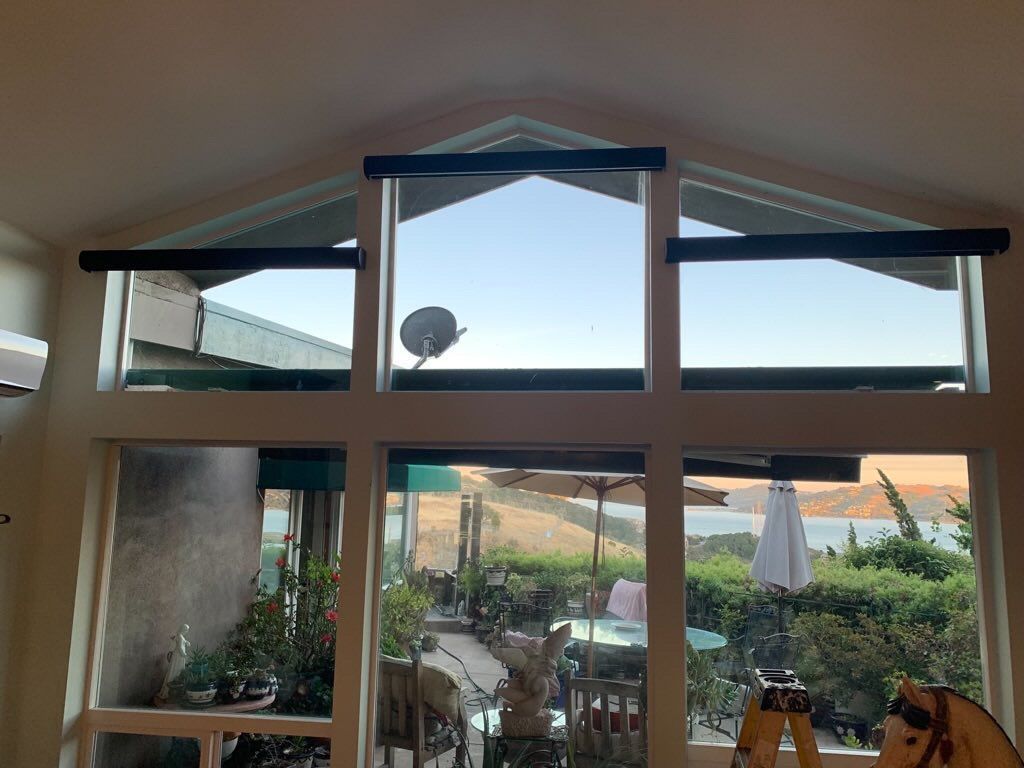 A large, multi-pane window with dark roller shades, framing a view of a patio, foliage, and a distant body of water.