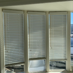 A bay window with three sets of white horizontal blinds partially drawn, showing a view of a street and cars outside.