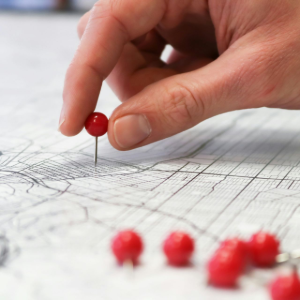 A hand placing a red pushpin on a detailed paper map, with several other red pins scattered on the surface nearby.